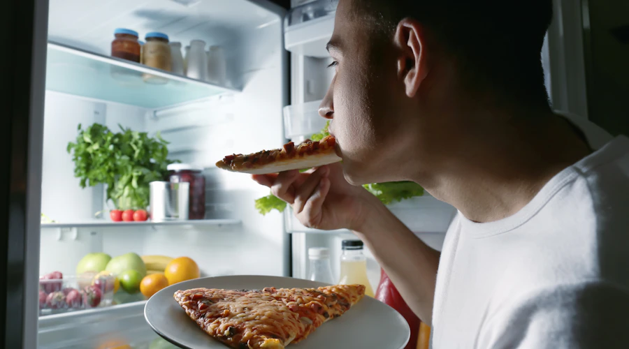 Man eating pizza directly out of the refrigerator at night