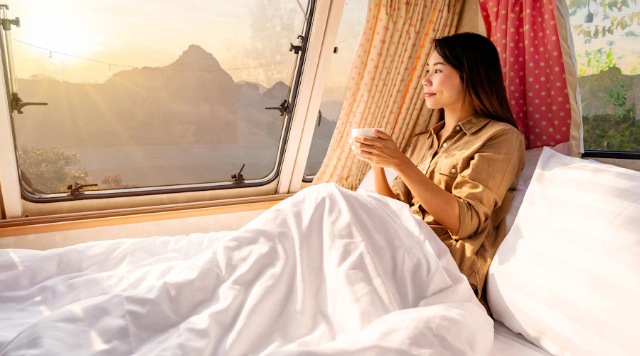 A woman drinks coffee in her camper bed with white sheets